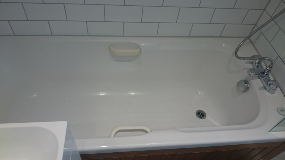 The image shows a clean and well-maintained bathroom bathroom with a white bathtub positioned against tiled walls. The bathtub has a built-in safety handle on one side and a silver centre drain. A modern silver faucet with a flexible hose and showerhead attachment is mounted on the right side, adjacent to a small chrome knob for water control. The surrounding tiles are light grey with a subway pattern, creating a contemporary look. The surface of the bathtub appears spotless, indicating it has been professionally cleaned, which is representative of services offered in Istead Rise (postcode DA10). The lighting in the room is bright, highlighting the shiny, polished finish of the tub and fixtures. Overall, the space is tidy, functional, and suitable for cleaning services aiming to maintain high hygiene standards in residential bathrooms, consistent with the offerings of Tidy Up in the local area.