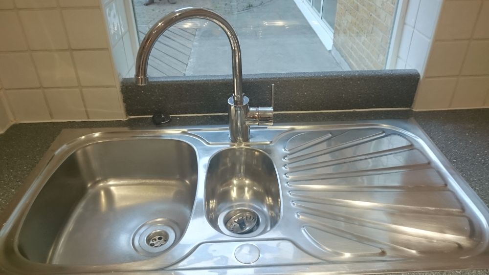 A stainless steel kitchen sink with two basins and a drainer, installed in a dark grey speckled countertop. The larger left basin appears clean and shiny, with a central drain. The smaller right basin also looks polished and free of debris. A modern chrome faucet with a curved design is mounted above the sink, with a single lever handle for controlling water flow. Behind the sink, there is a backsplash of beige tiles, and beyond that, a window with a view of a patch of outdoor decking and a brick wall, indicating the room is in a kitchen area. The overall setting is well-lit, emphasizing the cleanliness and polished surface of the sink and surrounding workspace, consistent with professional cleaning standards in Walton on Thames, KT12, managed by Tidy Up’s cleaning services.
