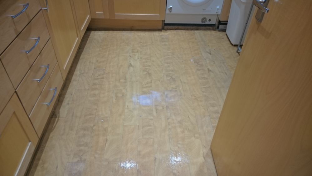 A view of a kitchen floor in a residential setting with a light-colored wooden laminate surface that appears clean and polished, reflecting some light. The flooring is bordered by wooden cabinets on the left side with silver handles, and the space includes a washing machine or dryer at the back. The kitchen area also contains visible appliances and a glimpse of a white refrigerator on the right side. The room is well-lit, highlighting the detailed wood grain of the cabinets and the glossy finish of the laminate flooring. The tidy and well-maintained appearance of the space suggests professional cleaning, consistent with services offered by companies like Tidy Up in Bexley, DA15, and nearby areas. The image underscores the importance of regular cleaning to keep kitchen surfaces, floors, and appliances in pristine condition.