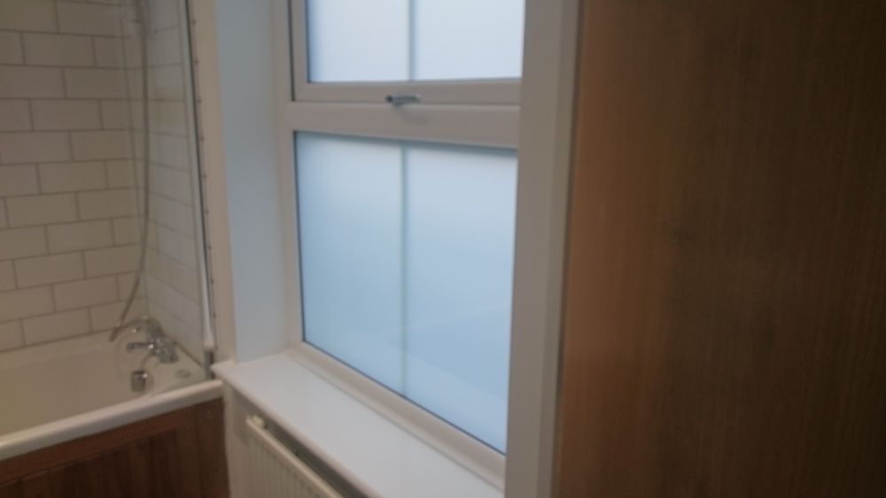The image shows a section of a bathroom, featuring a frosted glass window with a white frame situated above a radiator. To the left, there is a corner of a bathtub with a white interior and wooden paneling along its exterior, with part of a chrome showerhead and hose visible attached to a brick-patterned wall. The overall space appears clean and well-maintained, with the surfaces including the window ledge, radiator, and bathtub looking free of dirt or clutter. Natural light filters through the frosted glass, creating a soft, diffused atmosphere in the room. The wooden paneling around the bathtub adds a warm tone to the neutral colour scheme, complementing the white fixtures and light brick-patterned wall. This setting reflects a tidy, residential bathroom, likely one that [Tidy Up] might service for regular cleaning in Norbiton, KT2. The scene emphasizes the importance of maintaining clean, well-kept bathroom environments through professional cleaning services, highlighting aspects such as the cleanliness of surfaces, fixtures, and the overall neat presentation of the space.