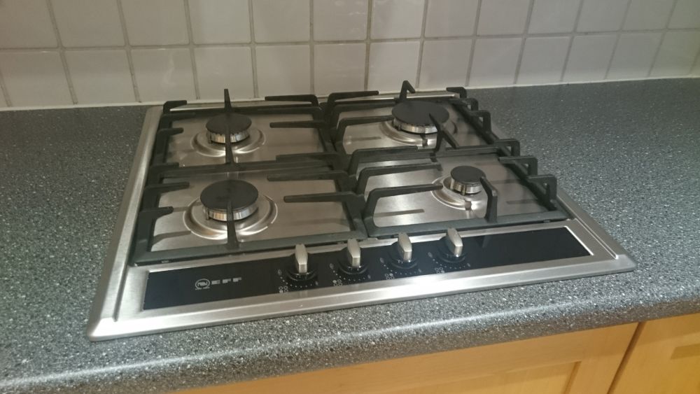 A stainless steel gas hob with four burners situated on a speckled grey kitchen countertop, set against a backdrop of beige tiled wall. The burners include a large front-left, front-right, back-left, and back-right burner, each with black cast iron pan supports. The control knobs are located centrally at the front of the hob, with a black surface and silver markings indicating heat levels. The countertop surface appears clean and free of debris, with no visible spills or stains, highlighting a well-maintained cooking area. The kitchen cabinetry below is made of light wood, and natural light illuminates the space, creating a bright and airy atmosphere. This setting exemplifies professional kitchen cleaning standards often offered by services such as Tidy Up for homes or commercial environments in Gravesend, DA11, Bay, in the UK, ensuring appliances are kept hygienic and presentable.