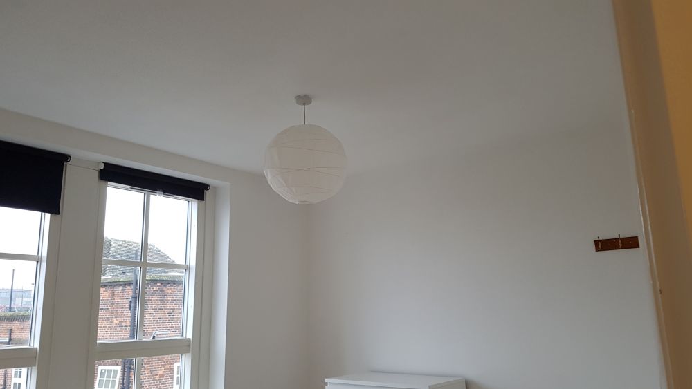 The image shows a clean and minimally furnished room with white walls and a white ceiling, featuring a round paper lantern ceiling light. The room has two large double-glazed sash windows with white frames, allowing natural light to brighten the space, and are fitted with black roller blinds, one of which is partially lowered. Outside the windows, neighboring brick buildings with pitched roofs are visible, indicating an urban residential area likely in Ashtead. The visible flooring appears to be light-colored and smooth, either wood or laminate, and the corner of a white piece of furniture is seen at the lower right. On the right wall, there is a small wooden hook or rail attached, suggesting a place for hanging items. The overall condition of the room appears pristine, with no visible dust or clutter, exemplifying professional cleaning standards, likely by Tidy Up, a local cleaning service. This simple yet well-maintained interior highlights a space that has been thoroughly cleaned and prepared, possibly after a service provided in the KT21 postcode area.