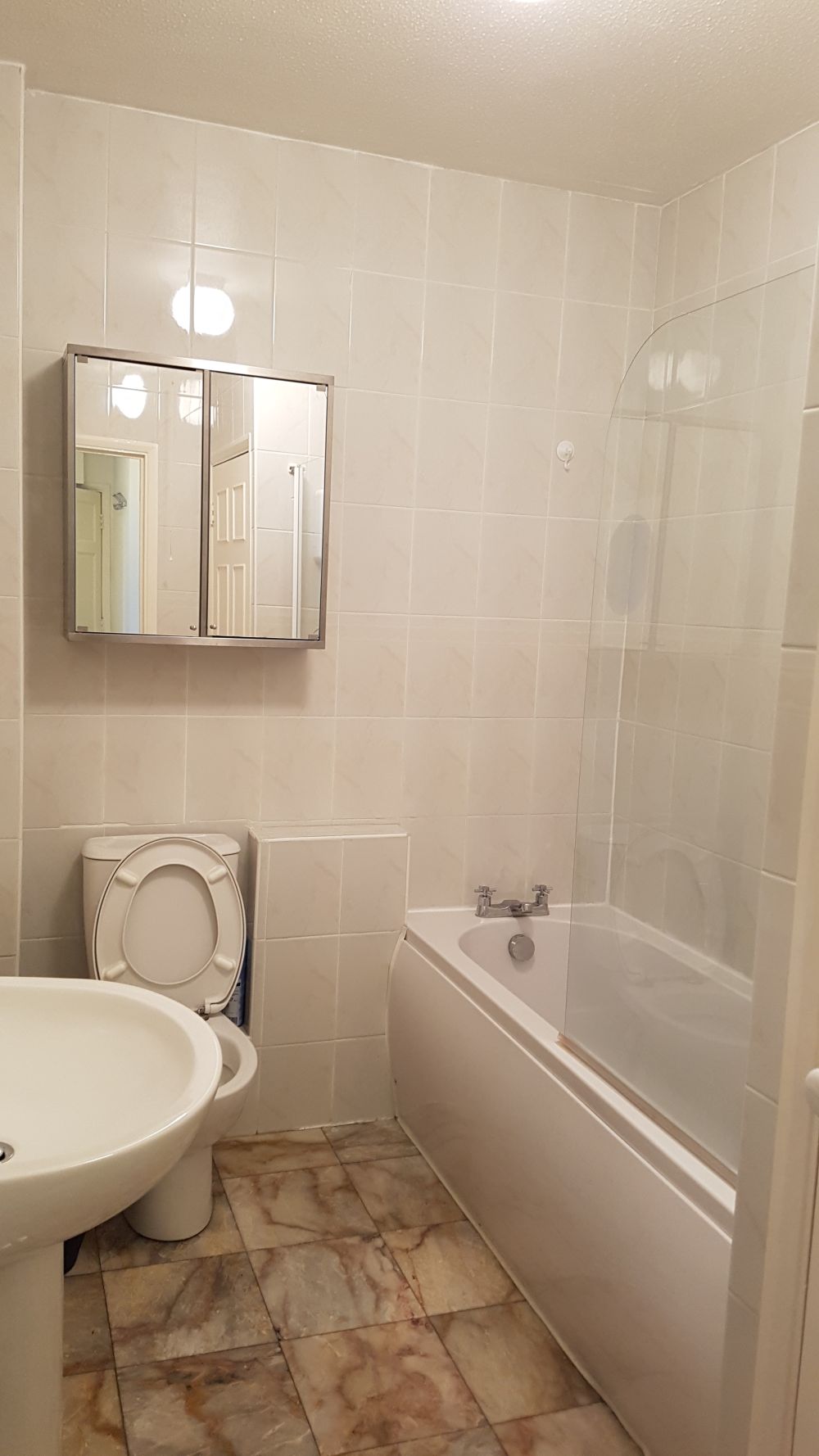 The image depicts a small, clean bathroom area with beige tiled walls and a patterned tiled floor. In the foreground, there is a white ceramic toilet with its lid open, positioned next to a partially visible white ceramic washbasin. Above the toilet, a mirror with a silver frame is mounted on the wall, reflecting a door and part of the hallway outside. To the right of the toilet, there is a white bathtub fitted with a transparent glass shower screen, which curves slightly outward. The bathtub appears to have a silver faucet with two handles. The lighting in the room is bright, creating a neutral and hygienic atmosphere. The overall condition of the surfaces suggests the bathroom has been recently cleaned, reflecting the professional cleaning services offered in West Thurrock, RM20, by Tidy Up, as part of their cleaning category.