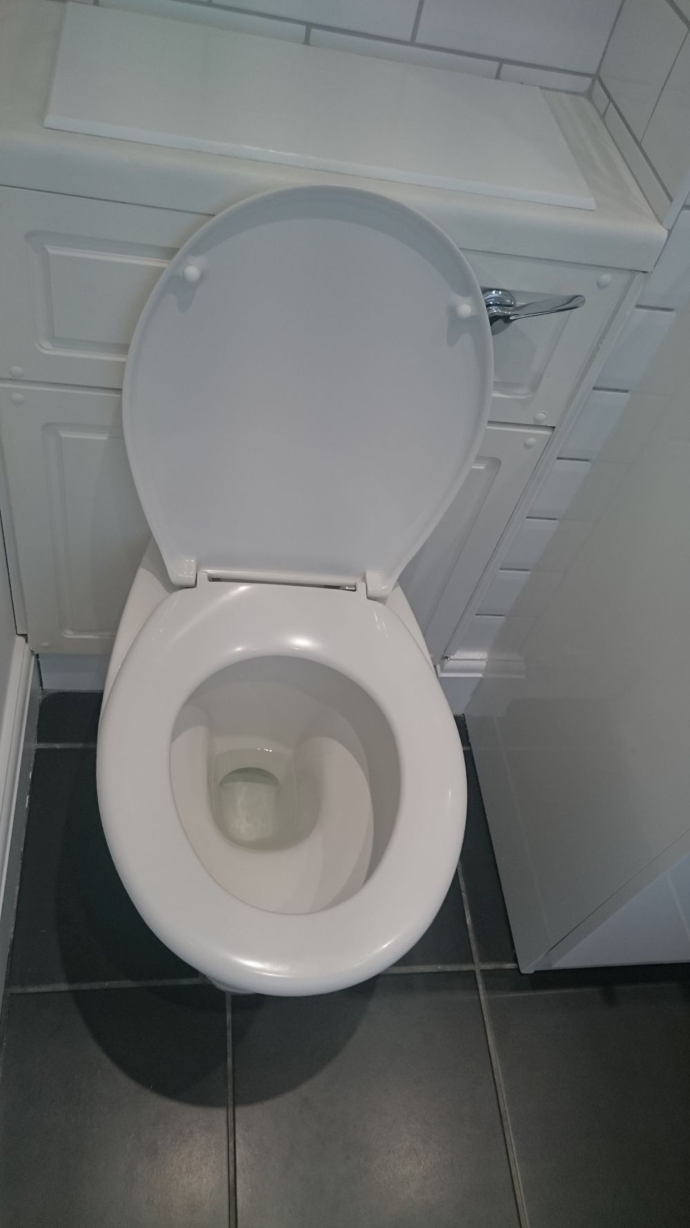 The image shows a clean, white ceramic toilet with an open lid in a small bathroom area. The toilet is positioned against a wall with white tiled surfaces, reflecting a tidy and well-maintained space. To the right of the toilet, there is a white base cabinet with a visible chrome handle, likely part of the bathroom storage or sink unit. The floor is covered with dark grey tiles, which contrast with the white fixtures and enhance the neat appearance of the area. The lighting is bright, emphasizing the cleanliness of the surfaces. The overall scene suggests a bathroom that has been professionally cleaned, aligning with services offered by Tidy Up in West Thurrock, RM20. The visible fixtures and layout indicate a typical modern bathroom designed for functionality and hygiene, with surfaces free of stains or dirt, ready for occupancy or use.
