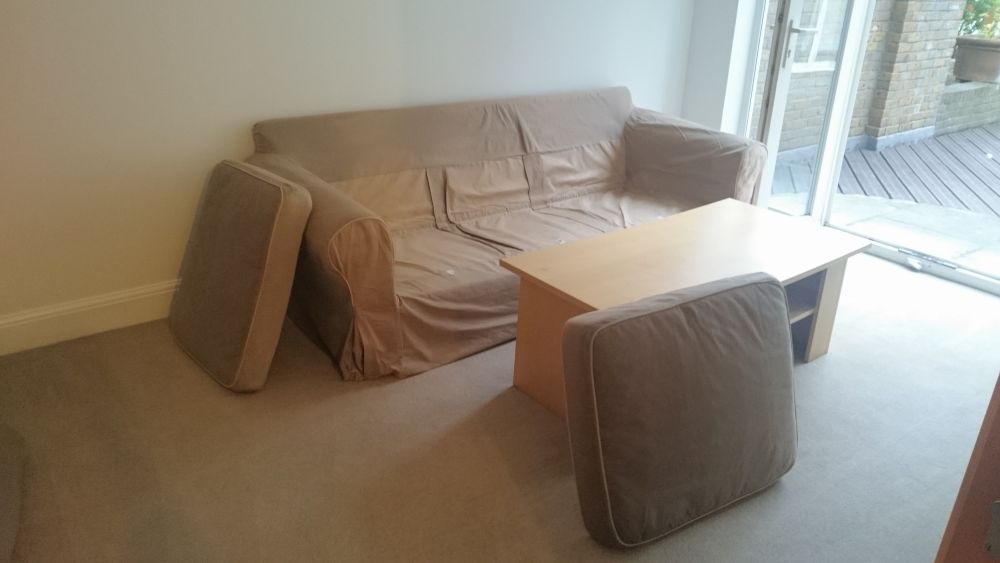 A living room area with a light beige fabric sofa positioned against a plain cream-colored wall. The sofa appears slightly wrinkled and has a loose cover or slipcover that is partially gathered at the sides. In front of the sofa, there is a small wooden coffee table with a light natural finish, placed directly on a cream carpeted floor. To the right of the sofa, a fabric armchair with matching beige upholstery is situated, with its back towards the viewer. The room is well-lit by natural light coming through large glass sliding doors on the right side, which lead to an outdoor wooden deck with a brick wall and a flower box visible outside. The overall space looks clean and tidy, with surfaces and textiles appearing well-maintained and free of visible dust or stains. The outdoor area adds a subtle contextual link to the Romford postcode, which is associated with the local cleaning service industry served by Tidy Up.