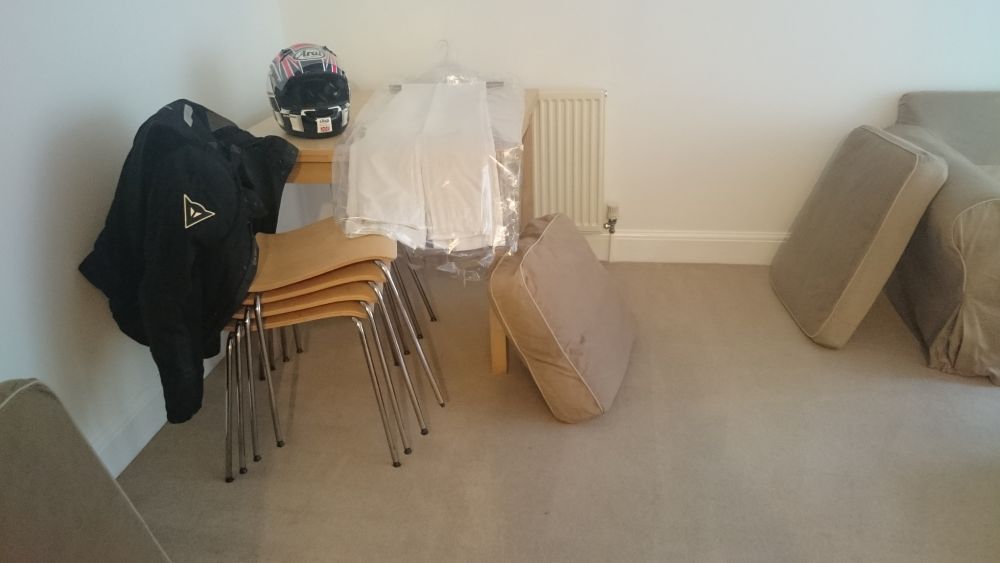 The image depicts a light-colored living room or lounge area with a beige carpeted floor. In the foreground, there are two beige cushions or small mattresses placed on the floor. To the right, part of a matching beige sofa is visible. In the background, against a white wall, there is a radiator with a white cover. Near the radiator, a small wooden side table is situated, supporting a clear plastic bag containing folded white towels or cloths, topped with a black and red motorcycle helmet. Adjacent to the table, on the left and partially overlapping it, are four stacked beige chairs with metal legs, and hanging over the chairs is a black jacket. Also on the table, a black backpack is positioned. The overall scene suggests a tidy, well-maintained space being prepared or cleaned, with visible elements of furniture, textiles, and personal items, reflecting a typical Southall residential setting within the UB1 postcode area, relevant to professional cleaning services by Tidy Up.