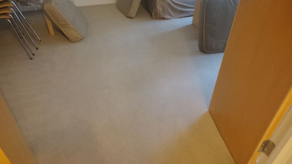 The image depicts a section of a room, most likely a living area or hallway, with a beige carpeted floor that appears clean and well-maintained. In the foreground, a wooden door with a brass hinge is partially visible, leading into the room. To the left, there is a small area with stacked metal chairs, suggesting a versatile or communal space, and a beige bag resting against the wall. Towards the back of the room, there are two armchairs upholstered in grey fabric, positioned with one slightly behind the other, indicating a conversational or relaxation area. The lighting is natural, coming from an unseen window, casting soft shadows across the neutral-toned carpet. The overall scene suggests a tidy, orderly space that could benefit from professional cleaning services like those offered by Tidy Up in Hertford, ensuring the surfaces remain spotless and inviting for residents or visitors. The setting is characteristic of a typical UK home, with practical furnishings and a focus on cleanliness and comfort, subtly supporting local SEO for Hertford cleaning services.