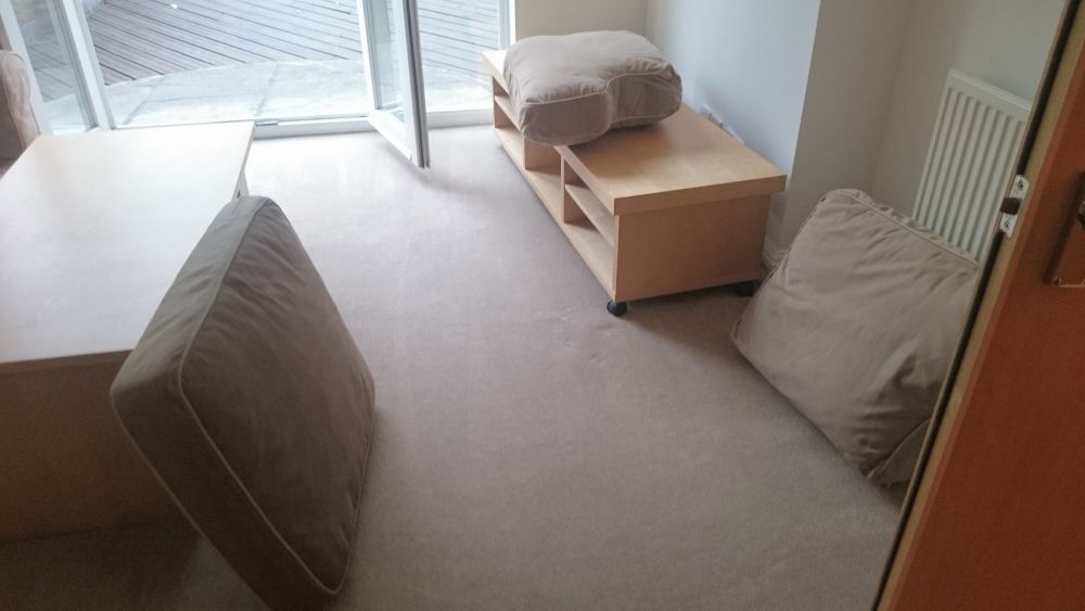 A photograph of a clean, minimalist living room with beige carpet flooring. In the foreground, part of a beige upholstered armchair is visible on the left. To the right, a second similar armchair is placed against the wall, with a large beige cushion resting on the floor beside it. Against the wall near the window, there is a light wood side table with open shelves, topped with a matching beige cushion. The room features a large glass sliding door with a view of a wooden deck outside, allowing natural light to brighten the space. The interior has white walls, and a white radiator is mounted on the wall next to the door. The overall arrangement and condition of the room suggest it has been thoroughly cleaned, aligning with professional cleaning services offered by Tidy Up in Romford (RM1 postcode).