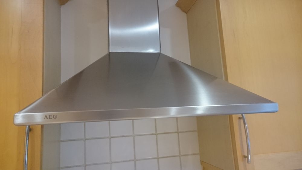 The image shows a close-up view of a stainless steel kitchen extractor hood mounted above a tiled cooking area in a modern kitchen setting. The hood has a smooth, polished surface with a slight professional sheen, and features the brand name 'AEG' inscribed on the front edge. The background includes cream-white tiled backsplash tiles arranged in a grid pattern, with visible grout lines, and light wooden cabinetry on either side of the hood, likely part of a contemporary kitchen design. The lighting appears natural, illuminating the surfaces evenly, highlighting the cleanliness and well-maintained condition of the appliances and surrounding surfaces. This setup exemplifies modern kitchen standards, making it suitable for professional cleaning services specializing in kitchen appliances, ensuring optimal hygiene and shine for spaces such as those in Footscray, DA14, and surrounding areas.