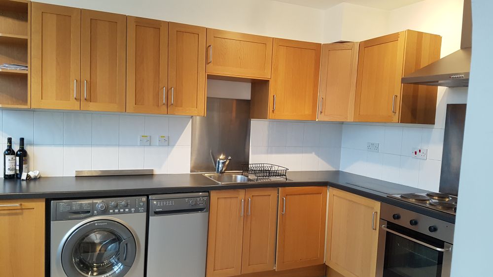A tidy kitchen in Windsor with wooden upper and lower cabinets, some of which have silver handles, and a dark grey countertop. The white tiled backsplash runs along the wall behind the counter, with two power outlets visible. On the left side of the counter, there are two bottles of wine and a small stack of papers. The central area features a stainless steel sink with a silver faucet, above which a minimalist open shelf shelf is visible. To the right of the sink, there is a black dish rack. Below the countertop, part of a washing machine and a dishwasher are visible, both with metallic finishes. On the far right, an electric cooker with four burners is situated next to a stainless steel extractor hood. The area appears clean and well-maintained, with natural light illuminating the surfaces, making the space look welcoming and suitable for professional cleaning services in TW18, Windsor. The overall scene reflects a typical, orderly kitchen ready for cleaning or maintenance, with a focus on functionality and neatness.