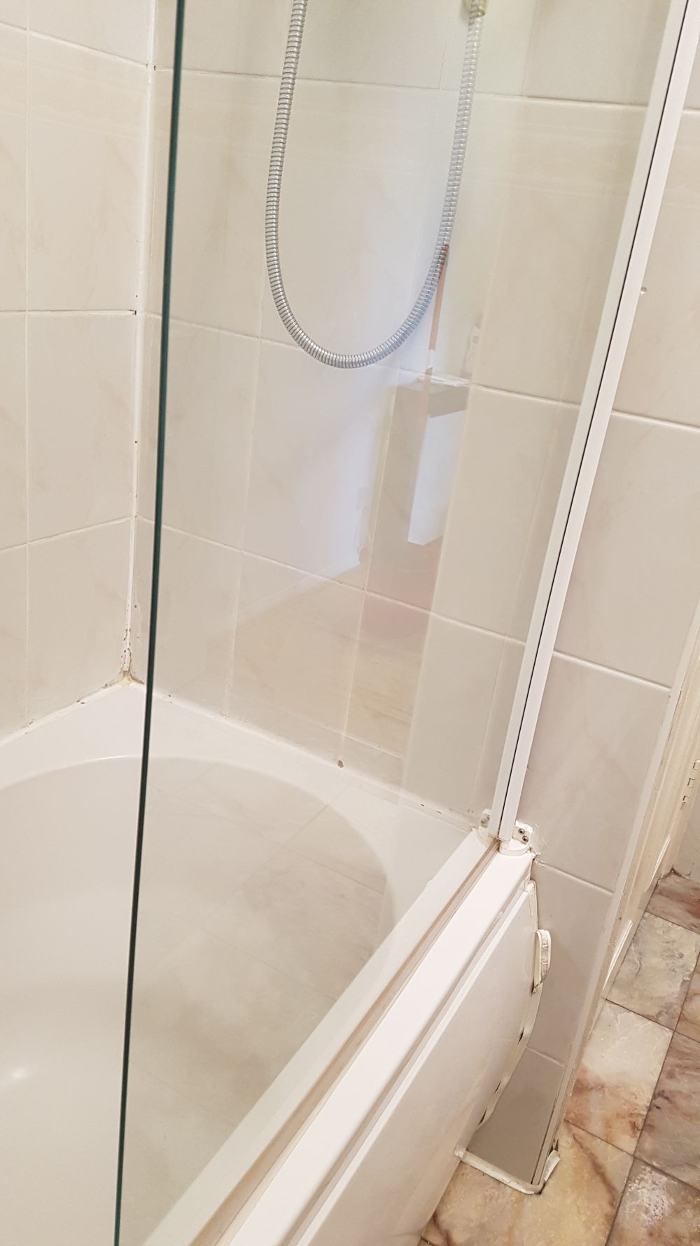 A close-up view of a shower enclosure in a bathroom located in Welwyn Garden City, with beige ceramic tiles on the walls and floor. The glass shower door is partially visible on the right side, with a white plastic frame and a handle. Inside the shower, a silver flexible shower hose with a showerhead attached is hanging, and a small white shelf with a bar of soap is mounted on the tiled wall. The floor is clean, and the overall area appears well-maintained, reflecting professional cleaning standards often provided by companies like Tidy Up in the AL8 postcode area.
