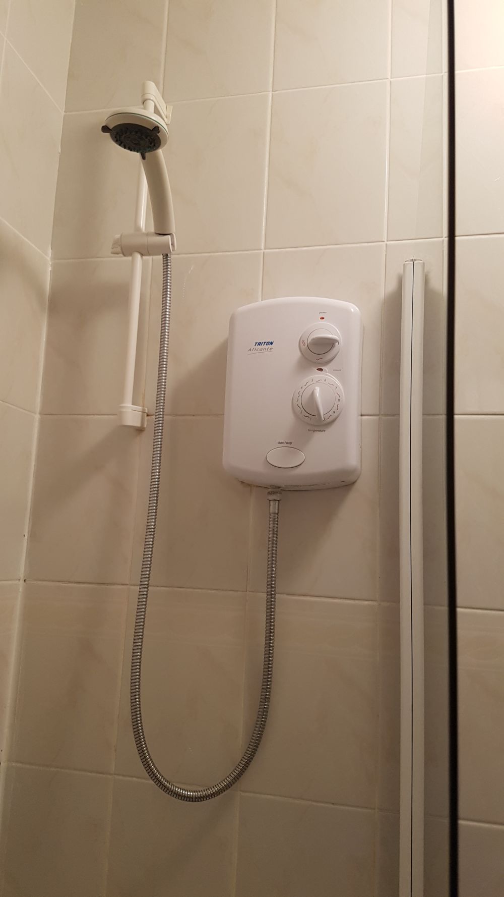 A close-up view of a bathroom shower area featuring beige ceramic tiled walls and a wall-mounted electric shower unit by Triton, with control knobs for temperature and power; the showerhead and adjustable hose are visible, with a vertical chrome rail securing the showerhead in place. The tiles are smooth and clean, reflecting bright lighting typical of a residential bathroom in West Drayton, UB7. The overall scene emphasizes the maintained condition of the shower space, suitable for professional cleaning services by Tidy Up that specialise in bathroom and kitchen cleaning in the local area. The image captures a neat, well-kept shower with clear visibility of surfaces and fixtures, ideal for maintaining hygiene and appearance in domestic environments.