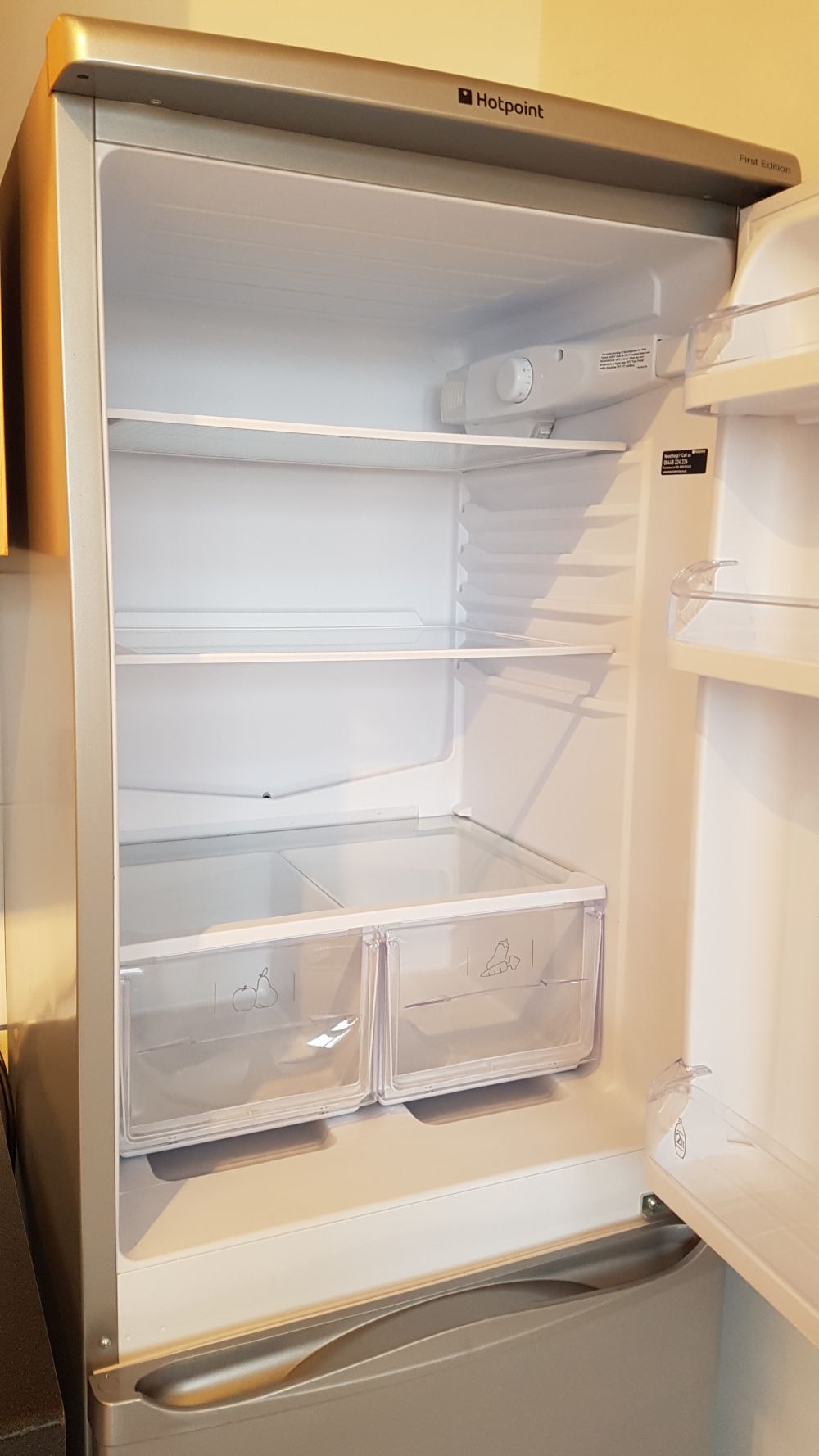 The image depicts the interior of an open, clean, and empty fridge in a kitchen, with white interior walls and shelves. The fridge has three main shelving levels, with the top shelf empty, the middle shelf also vacant, and the lower section featuring two clear plastic drawers, each illustrated with simple icons—one with a cherry and the other with a chicken leg—indicating storage for fruits and meats or poultry. The fridge’s interior appears well-maintained, free of stains or clutter, and the shelves are spotless. The overall setting suggests a modern, well-kept kitchen environment, with the fridge positioned against a light-colored wall. Subtle natural or artificial lighting illuminates the space evenly, highlighting the clean surfaces typical of professional cleaning service standards. The image subtly supports local SEO signals by being representative of a typical home kitchen in Chertsey with a focus on cleanliness, aligning with the services provided by Tidy Up, a cleaning company operating in the KT16 postcode area, specifically relevant to the kitchen appliances and interiors they clean.