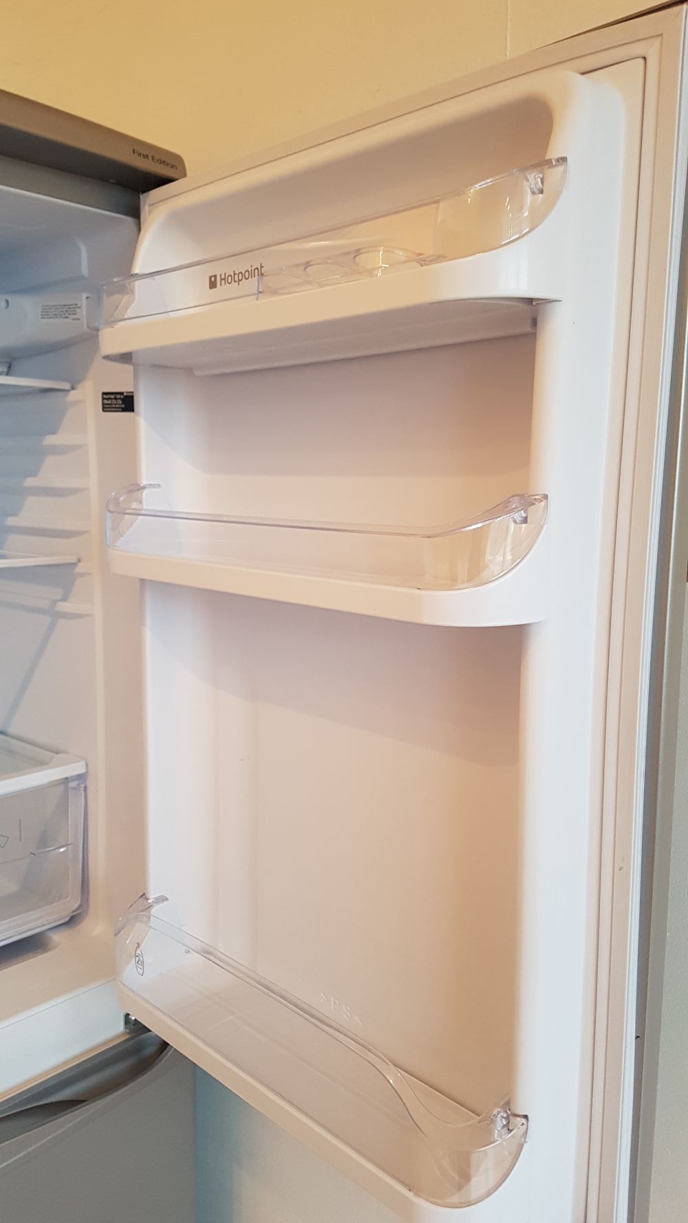 A close-up view of the door interior of a white household refrigerator, displaying three clear plastic door shelves. The top shelf has a slightly curved front edge and five small circular indentations, while the middle and bottom shelves are wider with a similar design. The shelves appear clean and unused, with no items stored on them. The background includes part of the fridge's interior with a glimpse of a drawer and door compartments, all in white plastic. The overall scene suggests a well-maintained kitchen environment. This image relates to cleaning services in Bracknell Forest, particularly for maintaining kitchen appliances. The lighting is neutral, highlighting the smooth surfaces and clarity of the plastic shelves in a typical domestic setting.