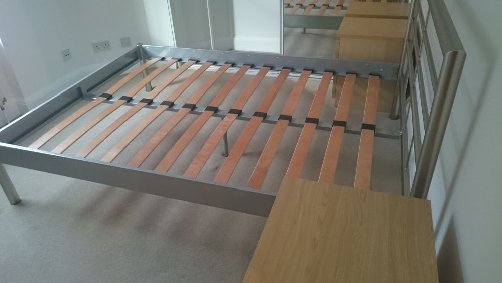 The image shows a metal bed frame with wooden slats positioned inside a room, next to a light-colored wooden table and a chair. The bed frame is placed on a carpeted floor near a white wall with multiple power outlets and switches. In the background, there is a large glass door or window leading to another room with wooden flooring and furniture, creating a bright and clean atmosphere. The metal frame appears to be newly assembled or awaiting assembly, and the room is well-lit with natural light. The setting indicates a typical residential bedroom, with visible surfaces that are clean and free of clutter, suggesting that professional cleaning services by Tidy Up in Cambridge could ensure the space remains tidy and pristine, especially in preparation for or following furniture assembly or renovation work.