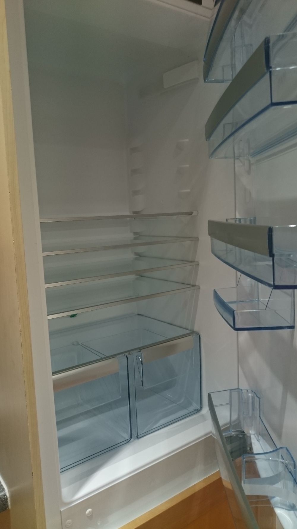 This image shows the interior of an open, empty refrigerator with white plastic shelves and transparent blue-tinted drawers. The refrigerator is clean and well-maintained, with multiple adjustable shelves and compartments visible, including a top shelf, middle shelves, and two clear drawers at the bottom. The interior light is on, illuminating the space evenly, highlighting the smooth surfaces and absence of stains or clutter. The refrigerator is positioned against a white kitchen wall, with a section of a wooden floor visible at the bottom. The overall scene suggests a recently cleaned or ready-to-clean appliance, consistent with professional cleaning services provided in residential kitchens within the Bracknell Forest area, specifically around SL4 postal code. The image demonstrates attention to detail with a focus on maintaining hygienic and visually appealing kitchen appliances, aligning with Tidy Up's professional cleaning standards.